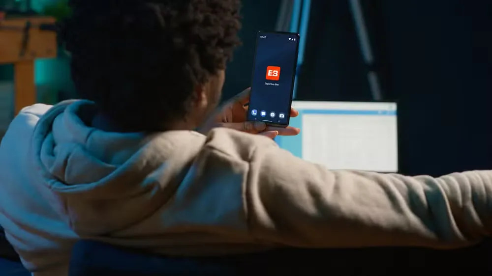 Pessoa de costas ao ar livre segurando o celular com um aplicativo de apostas na tela.